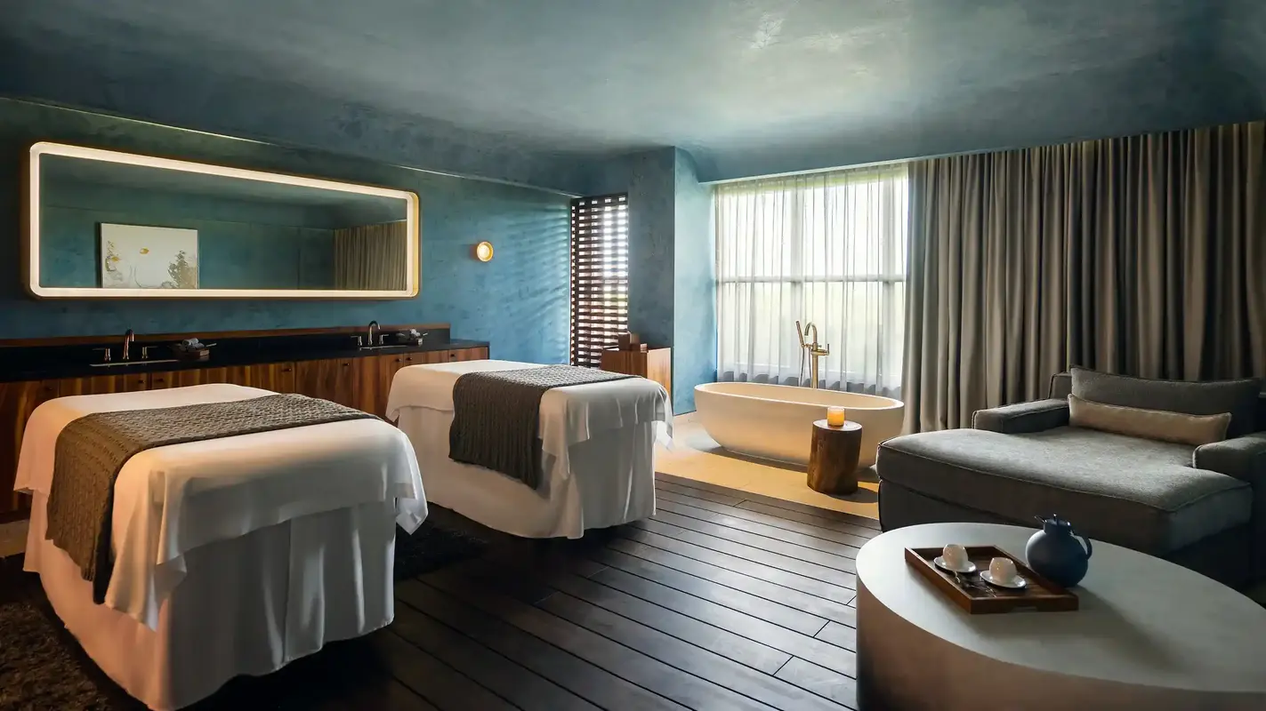 Spa treatment room with two massage tables, blue walls, soaking tub, and modern décor.